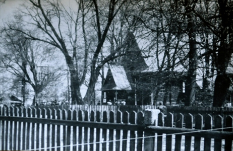 Otoczenie kościoła: naprzeciw głównego wejścia dzwonnica oraz parkan drewniany okalający teren przykościelny i teren cmentarza.